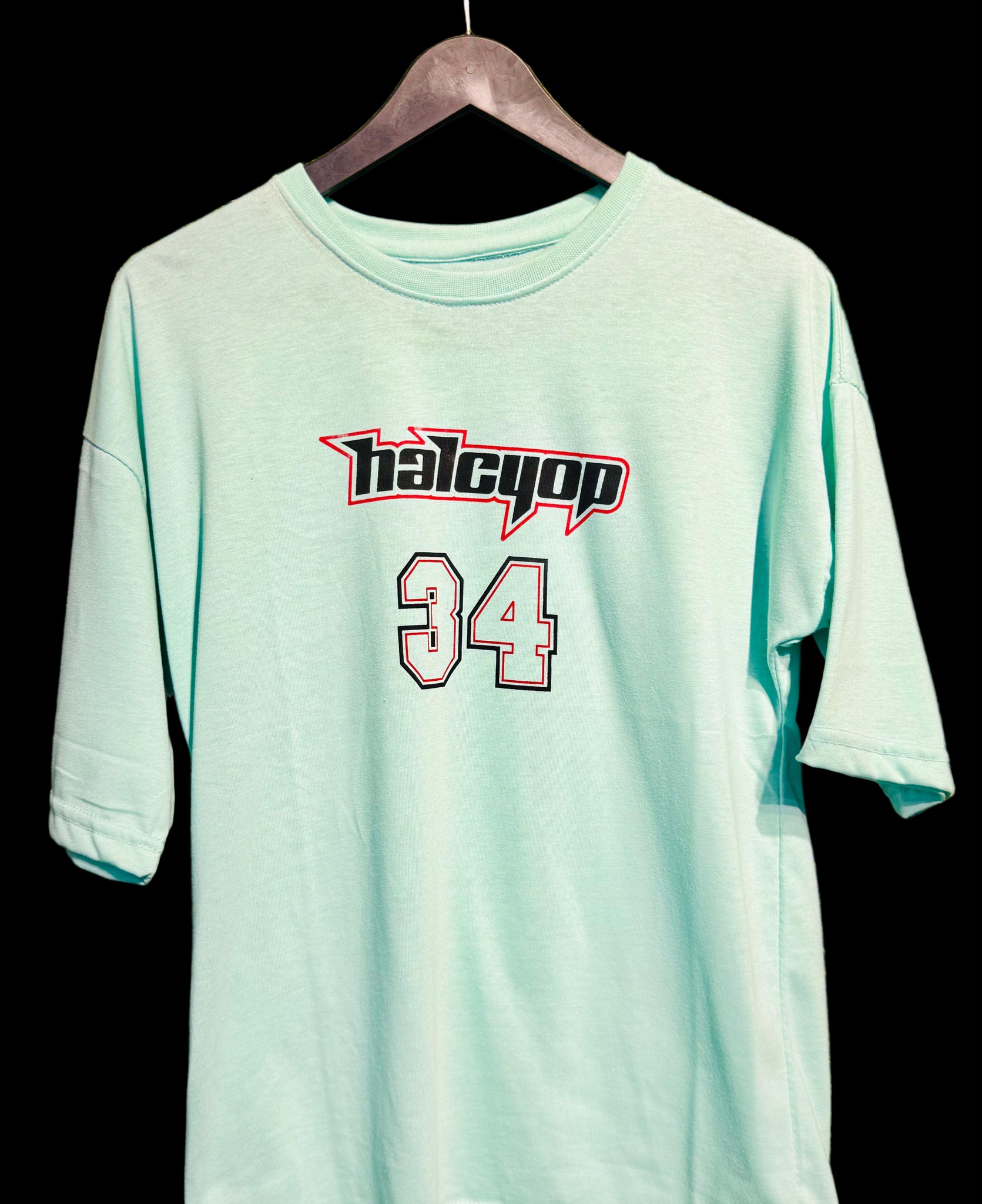 Light blue t-shirt with 'halcyon' and number '34' on a hanger against a green grass-like background